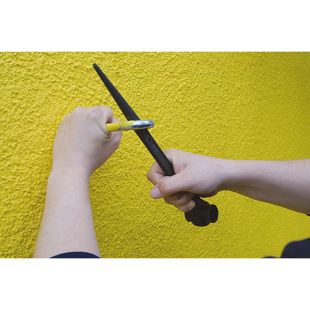 Hände halten einen schwarzen Stab mit einem gelben Werkzeug. Der Hintergrund ist eine gelbe Wand.